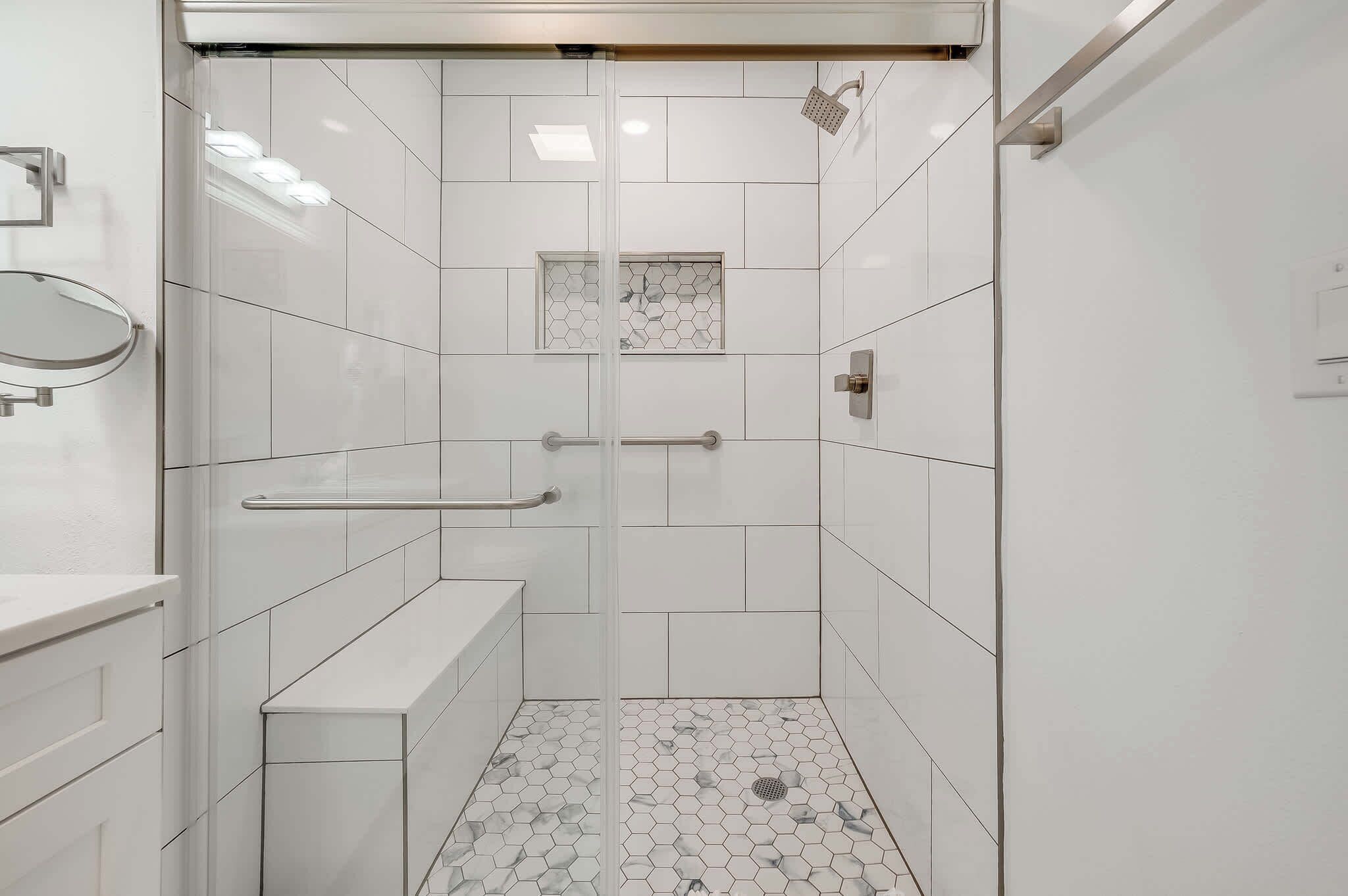 A modern bathroom with white tiles, a glass shower enclosure, a bench, and a hexagonal tile pattern on the floor.