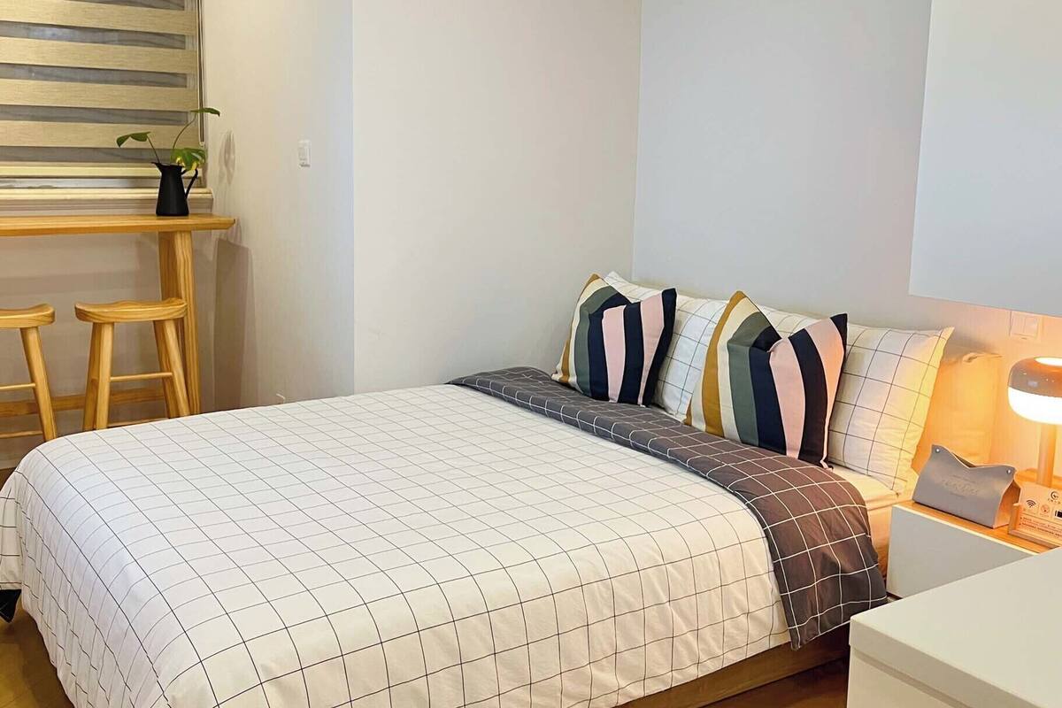 Quarto bem organizado com cama, mesa de cabeceira, abajur e um banquinho.