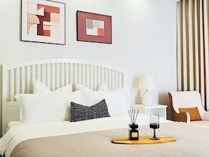Habitación de hotel con una cama blanca, una mesita de noche con una lámpara, una silla y dos obras de arte abstracto enmarcadas en la pared.