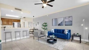 A modern living room with a blue sofa, a coffee table, and a dining area with a bar. - Trendy lake condo-fireworks view (Orlando)