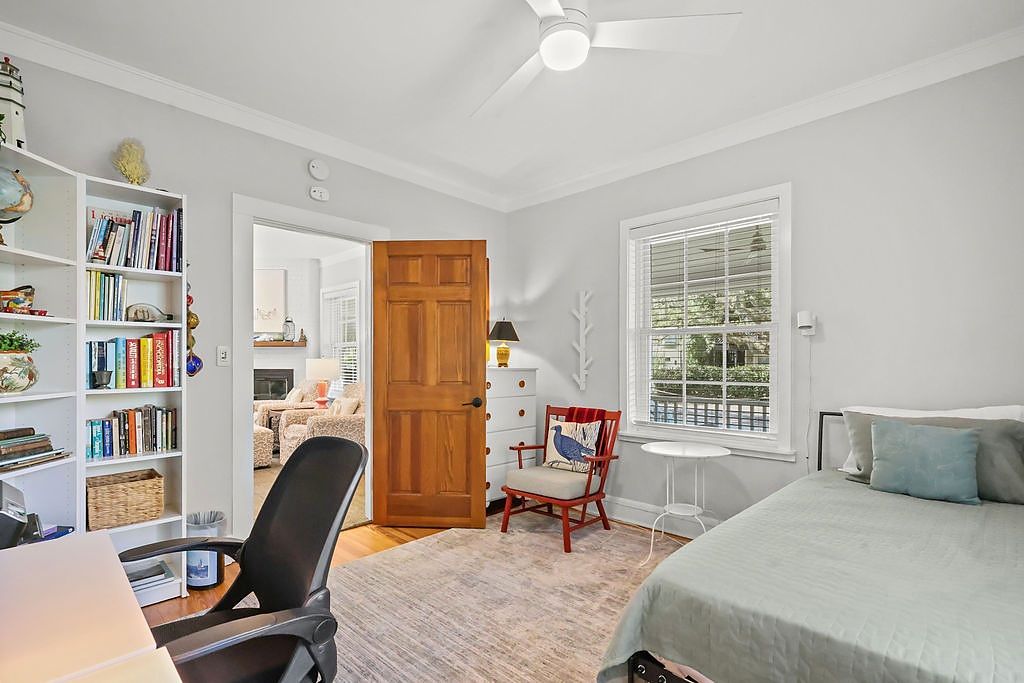 Une chambre à coucher avec un lit, un bureau avec une chaise, une bibliothèque, une porte en bois et une fenêtre avec des stores.
