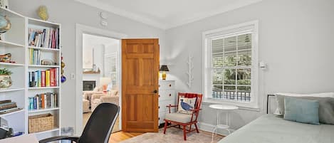 Une chambre à coucher avec un lit, un bureau avec une chaise, une bibliothèque, une porte en bois et une fenêtre avec des stores.