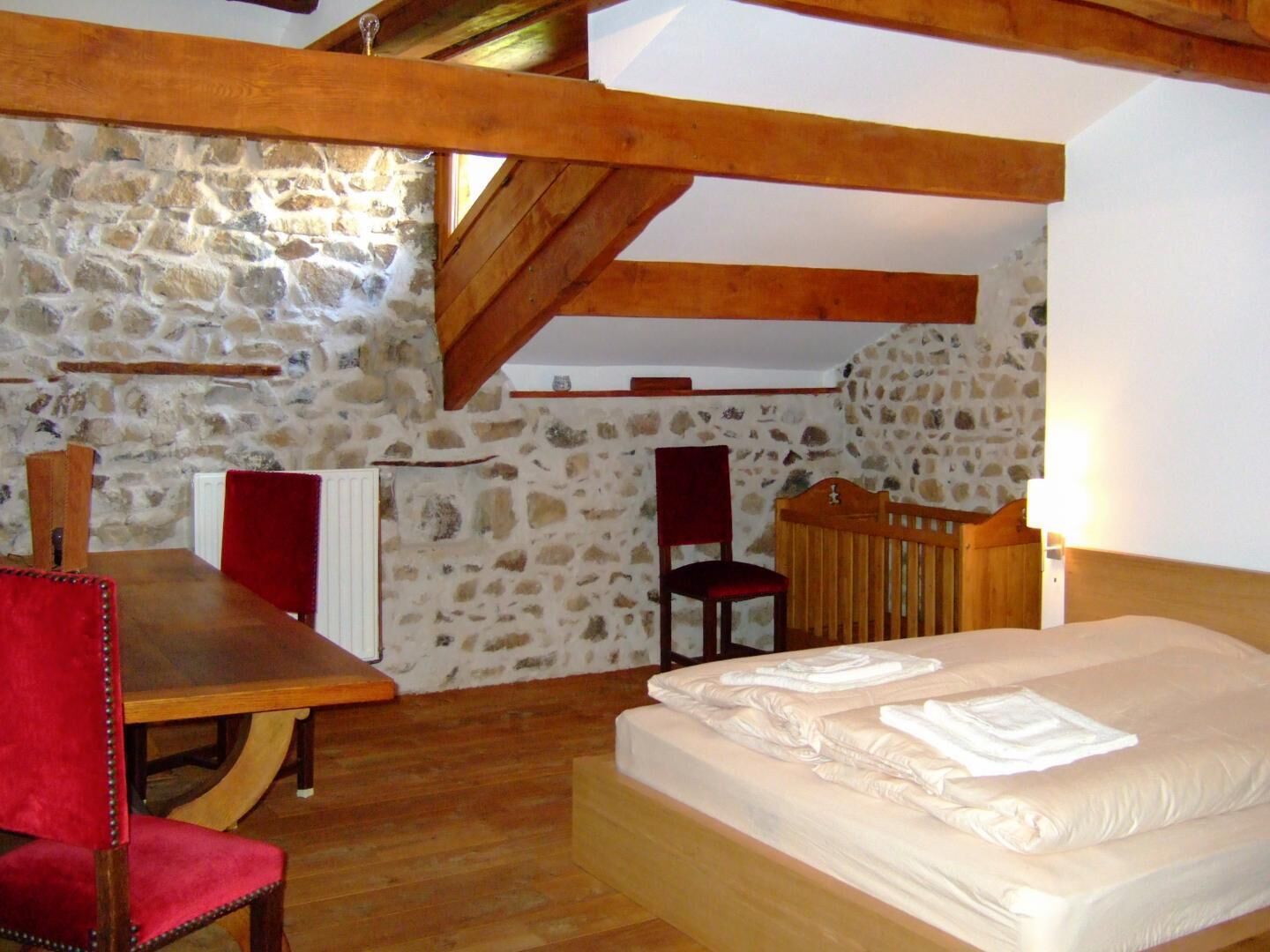 Une pièce chaleureuse avec un mur en pierre, des poutres apparentes, un lit avec du linge de maison blanc soigneusement plié, une table en bois et deux chaises rouges.