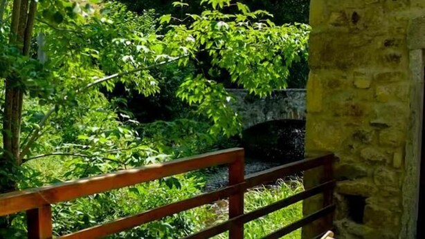 A wooden bench with a striped cushion in a sunlit outdoor area with a stone wall and lush greenery in the background. - Le Moulin Mathy - SAINT PIERRE LE VIEUX (SAINT PIERRE LE VIEUX)