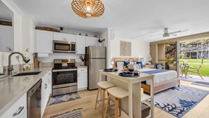 Une cuisine compacte dotée d’appareils électroménagers modernes, un coin repas avec des tabourets et une chambre avec un grand lit et vue sur le jardin.