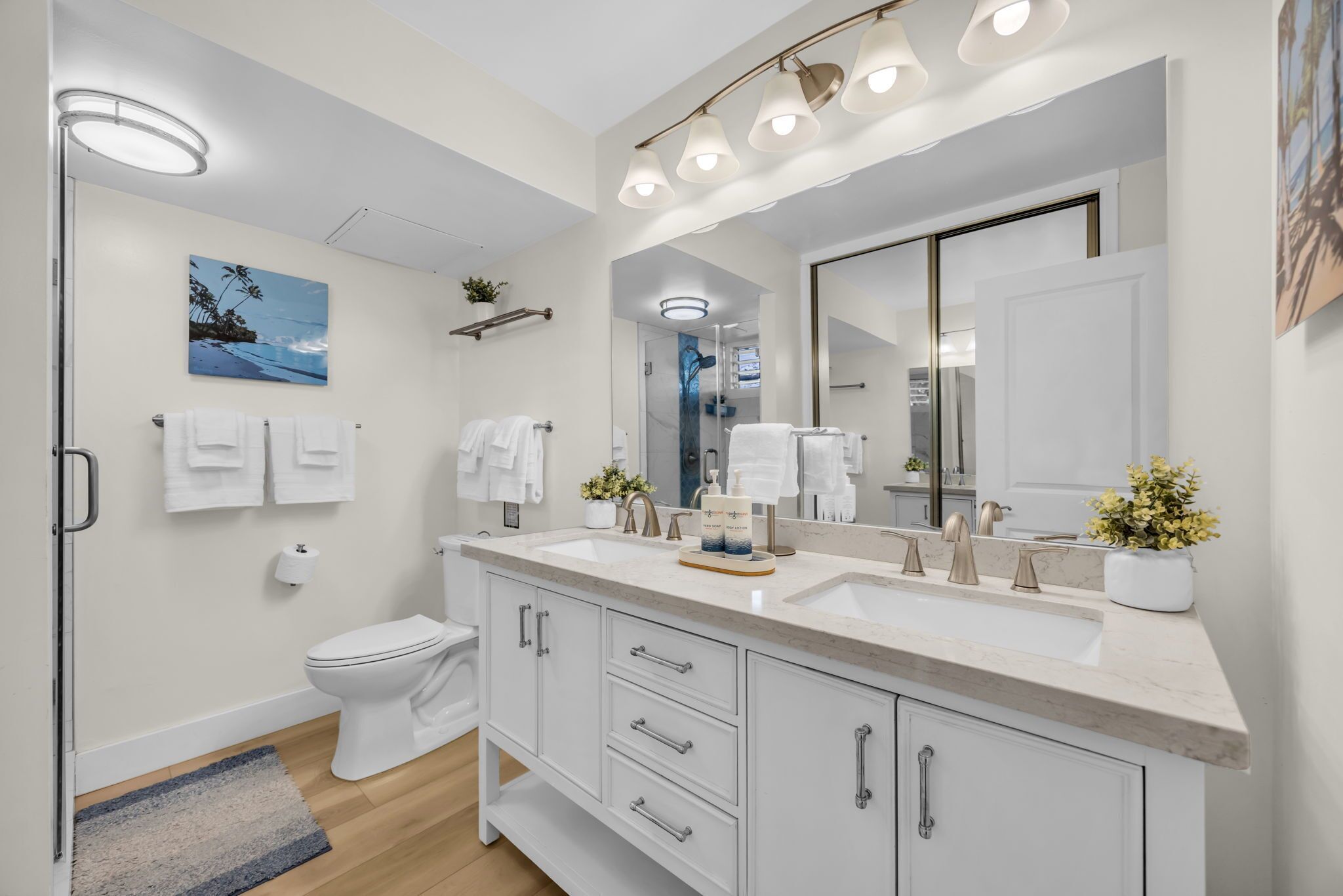 Une salle de bain moderne avec un grand miroir, deux lavabos et des toilettes. Des serviettes sont accrochées au mur et une petite plante est posée sur le comptoir.