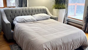 A neatly made bed with a grey upholstered headboard, a white pillow, and a grey bedspread. There is a large potted plant beside the bed, and a window with a view of trees and a wooden deck. - Charming Lakeview Cottage | Algonquin Shores (Dwight)