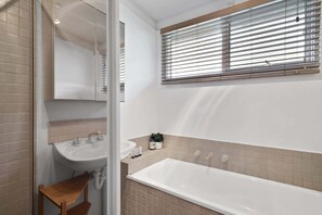 A bathroom with a bathtub, a sink, a mirror, and a window with blinds. - Casa Piccolo - Peninsula Hot Springs 5 mins away (Rye)