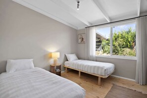 A bedroom with two beds, a bedside table with a lamp, a window with curtains, and a painting on the wall. - Casa Piccolo - Peninsula Hot Springs 5 mins away (Rye)