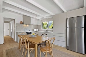 A modern kitchen with white cabinetry, a stainless steel refrigerator, and a dining area with wooden chairs and a table. - Casa Piccolo - Peninsula Hot Springs 5 mins away (Rye)