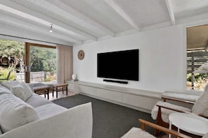 A modern living room with a large flat-screen TV mounted on the wall, a sofa, a small table, and a window with a view of greenery. - Casa Piccolo - Peninsula Hot Springs 5 mins away (Rye)