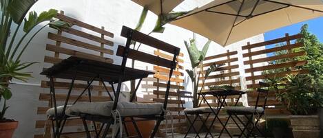 Un espacio al aire libre con bancos y mesas de madera, rodeado de plantas en macetas y una pared blanca.