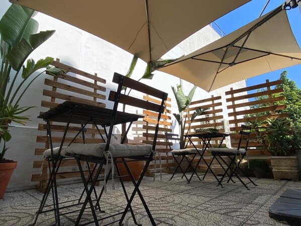 Un espacio al aire libre con bancos y mesas de madera, rodeado de plantas en macetas y una pared blanca.