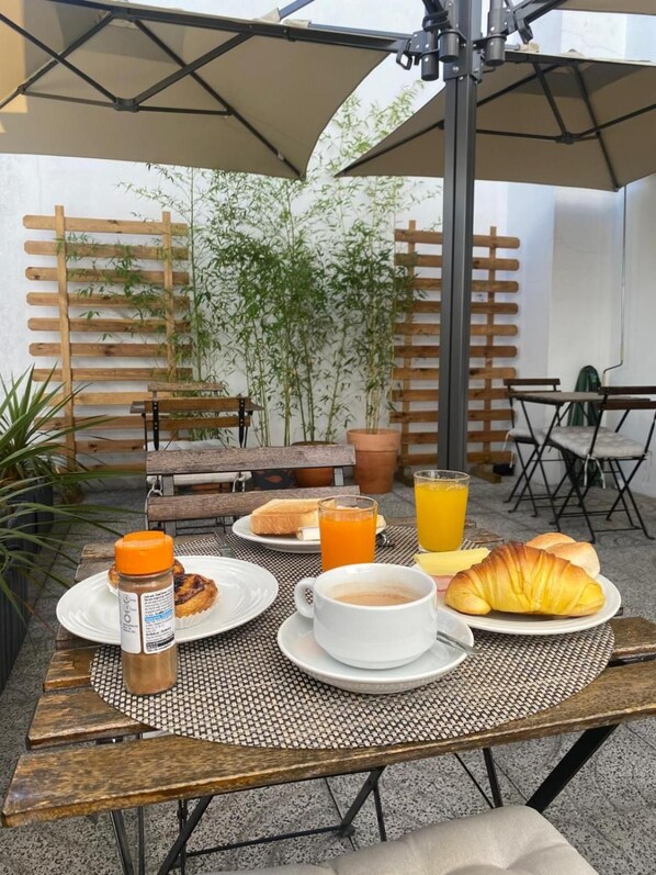 Un área de comedor al aire libre con una mesa dispuesta para el desayuno, que incluye cruasanes, tostadas y jugo, con un fondo de plantas de bambú y una valla de madera.