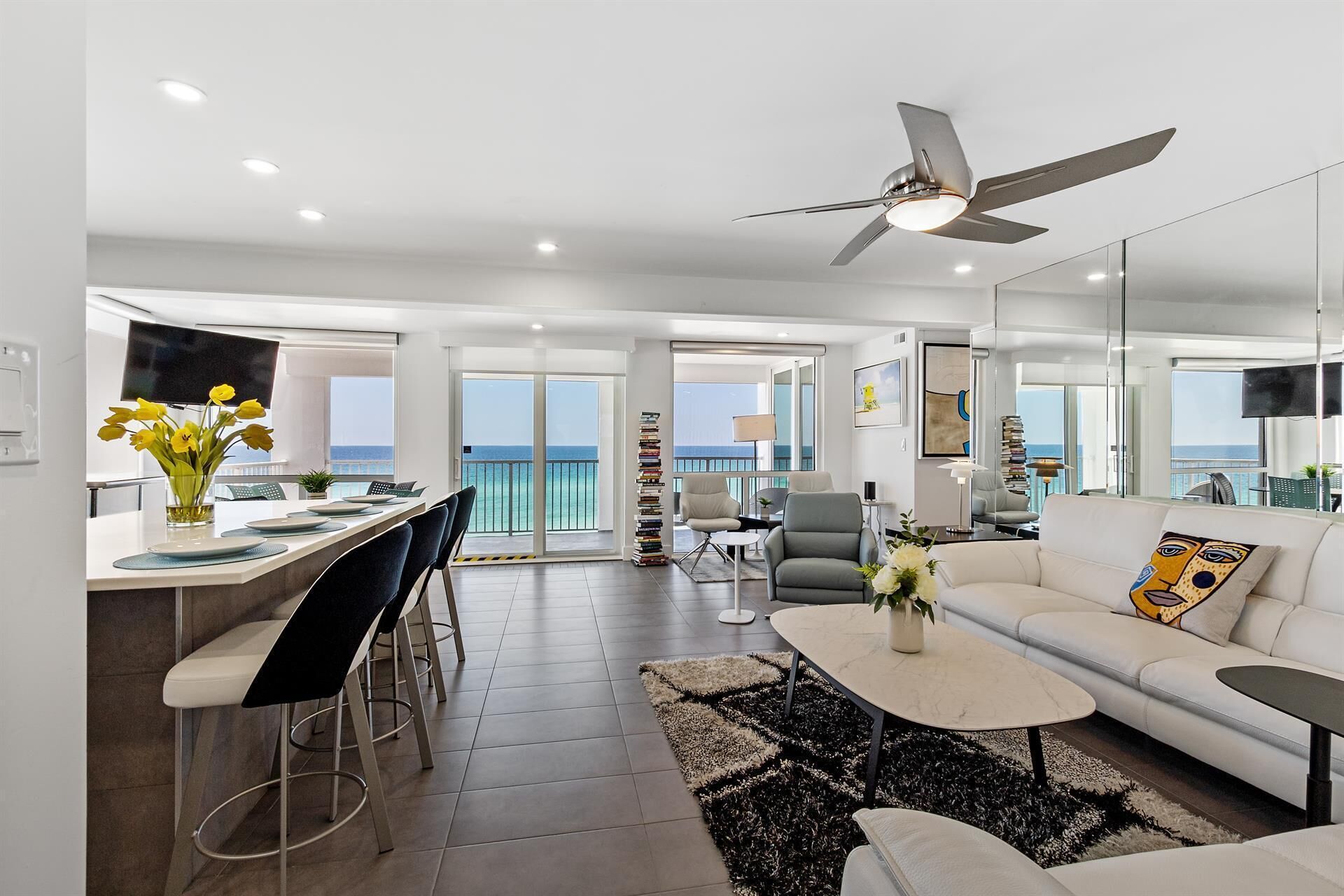 A modern living room with a sofa, coffee table, and a dining area with chairs and a small table. There is a ceiling fan, a TV, and a view of the sea through large windows.