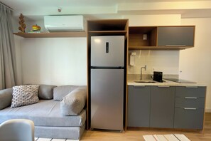 A compact living space with a sofa, a built-in refrigerator, a kitchen area with a countertop and cabinets, and a wall-mounted shelf with decorative items. - Apartment in Perdizes, 4th floor, with A/C, near Allianz Arena. (São Paulo)