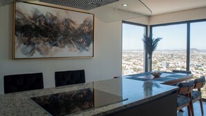 A modern kitchen with a large island, a dining area with chairs, and a large window offering a city view. - Sky & Sea View (Mazatlán)