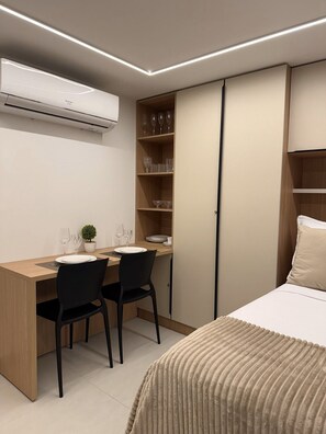 Quarto moderno com escrivaninha de madeira, duas cadeiras pretas, cama com manta bege, guarda-roupa e prateleira embutida com copos de vinho.