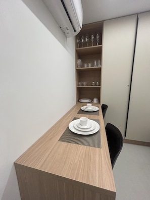 Ein gemütliches Hotelzimmer mit einem Holzespeisetisch, gedeckt für zwei Personen, einem kleinen schwarzen Stuhl und einer eingebauten Regallösung mit Gläsern und Geschirr.