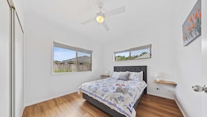 Quarto com cama, duas janelas, ventoinha no teto e piso de madeira.