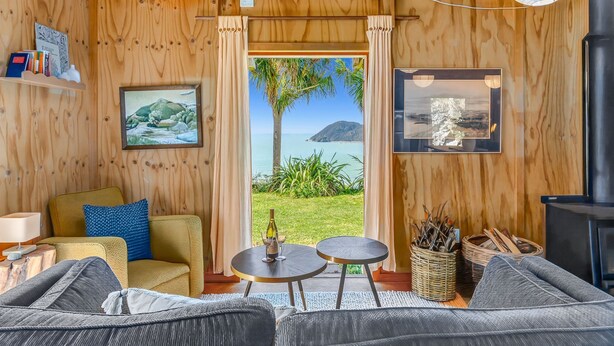 A wooden interior with a sofa, two small tables, a lamp, and a view of the outdoors. - The Gatehouse - Tata Headlands (Wainui Bay)