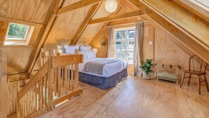 A wooden attic bedroom with a bed, a small seating area, and a window with curtains. - The Gatehouse - Tata Headlands (Wainui Bay)