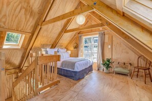 A wooden attic bedroom with a bed, a small seating area, and a window with curtains. - The Gatehouse - Tata Headlands (Wainui Bay)