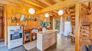 A wooden interior with a kitchen area featuring a stove, oven, and hanging pots. There is a wooden counter with a vase of flowers and a refrigerator in the background. - The Gatehouse - Tata Headlands (Wainui Bay)