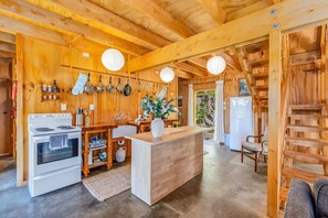 A wooden interior with a kitchen area featuring a stove, oven, and hanging pots. There is a wooden counter with a vase of flowers and a refrigerator in the background. - The Gatehouse - Tata Headlands (Wainui Bay)