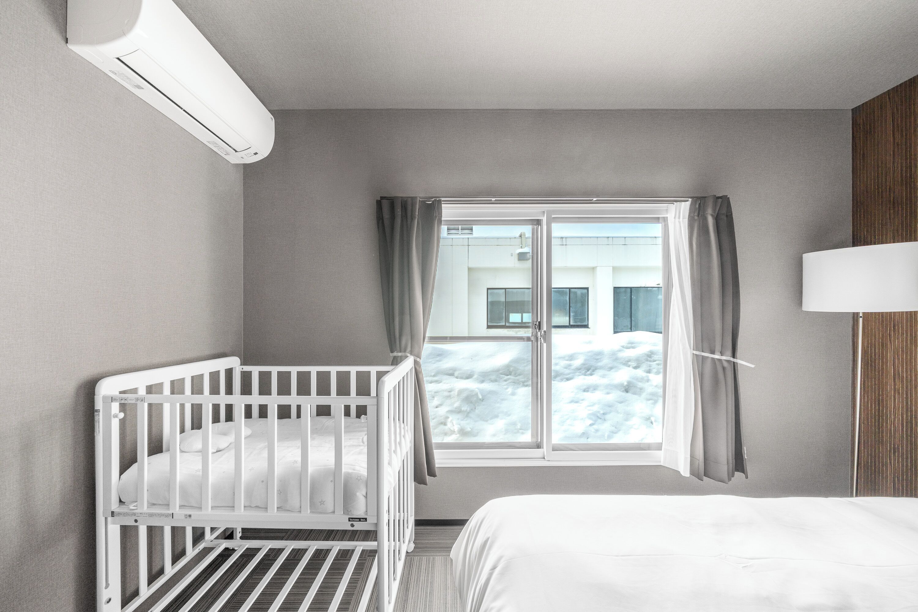 A hotel room with a crib, a bed, a window with curtains, and a wall-mounted air conditioner.
