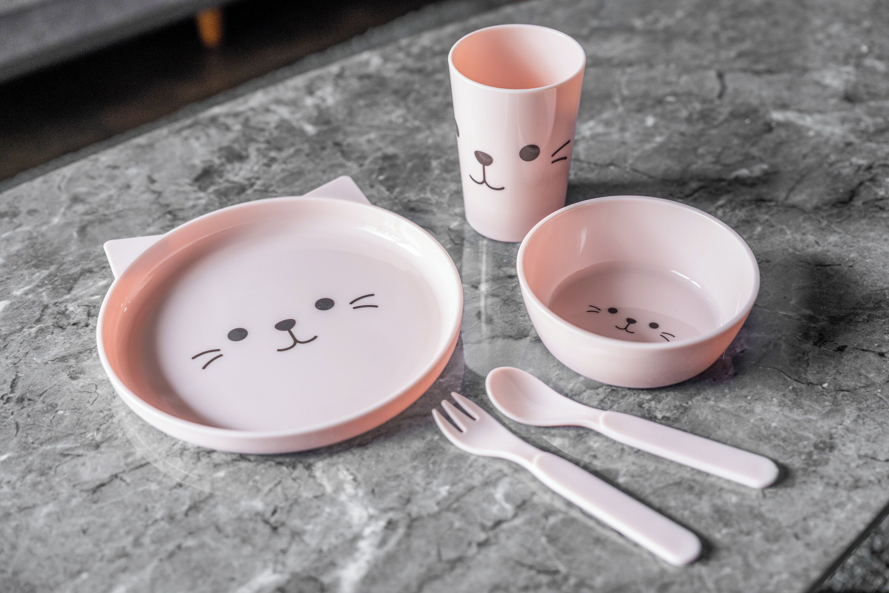 A set of pink ceramic dishes with cat faces, a pink cup, and a fork on a textured surface.