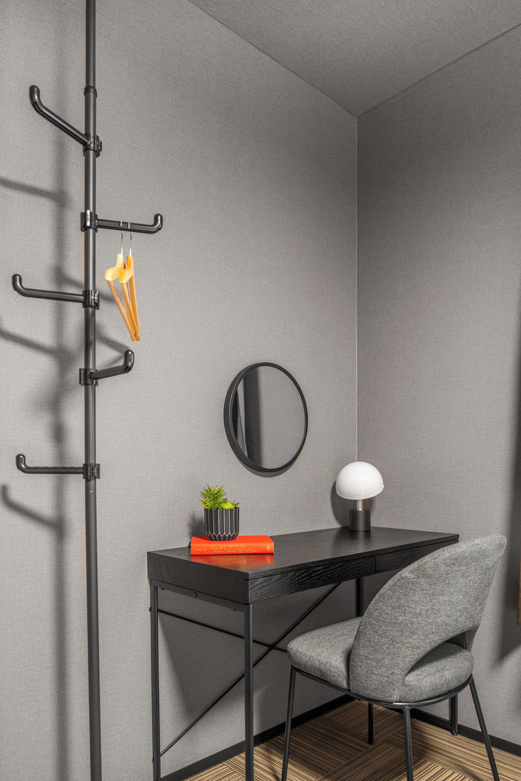 Une chambre moderne avec un bureau noir, une chaise grise, une lampe fixée au mur, un miroir et un porte-manteau sur lequel sont accrochés un manteau et une plante.