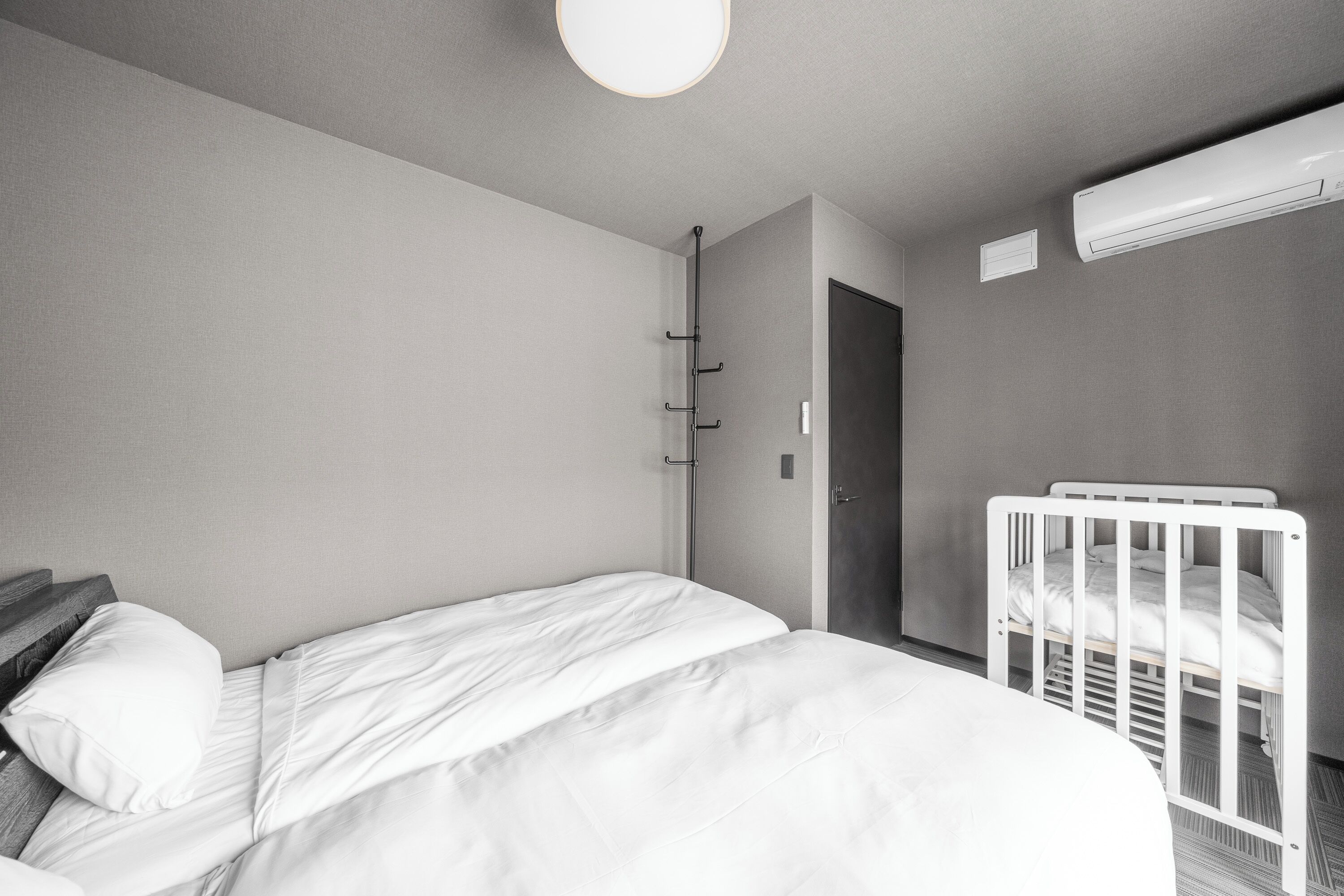 Habitación de hotel minimalista con una cama individual, una cuna blanca y un aire acondicionado instalado en la pared.