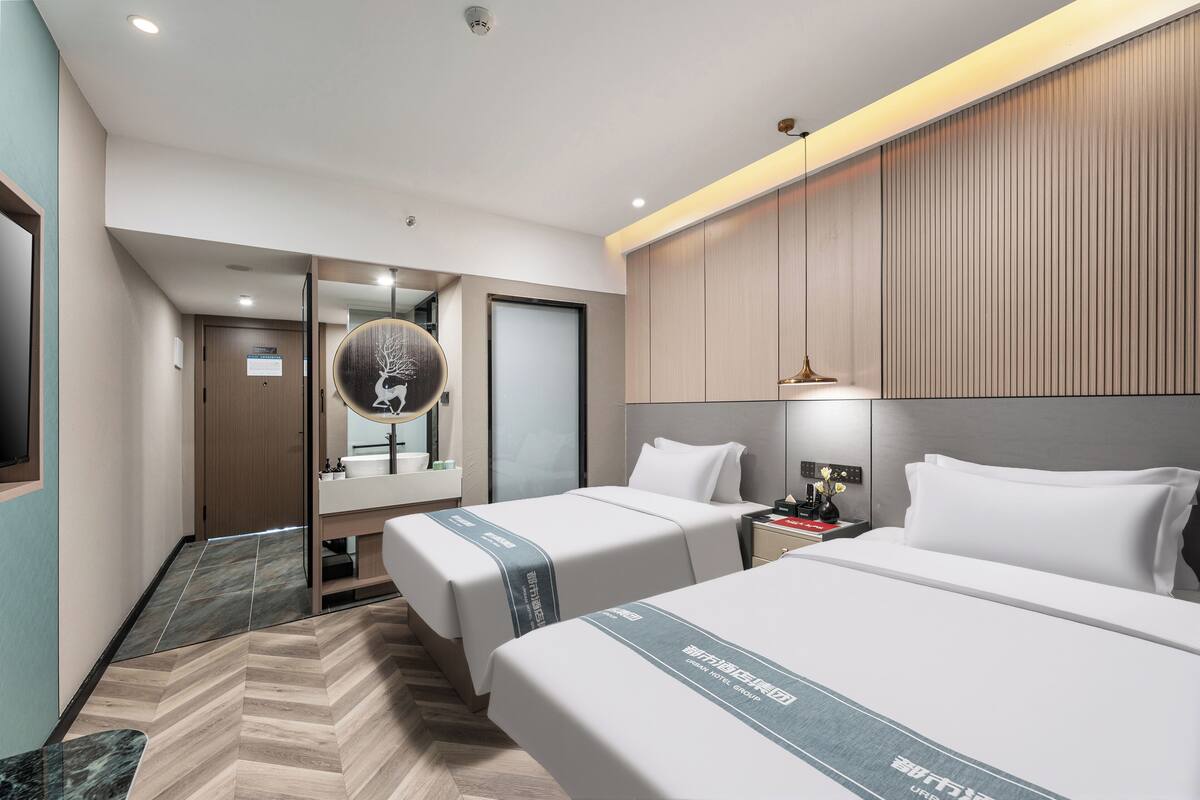 Habitación de hotel con dos camas, un espejo grande, una mesita de noche con lámpara y vista al pasillo.