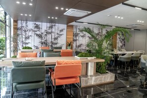 A modern dining area with a long table, orange and green chairs, and a large potted plant.