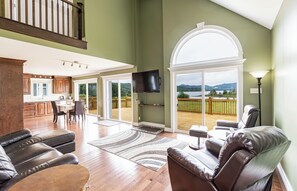 A spacious living room with a large window, a flat-screen TV mounted on the wall, and a dining area visible through the glass doors.