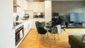 Moderni keittiö, jossa on valkoiset kaapit, ruokailutila vihreillä tuoleilla ja olohuoneessa litteänäyttöinen televisio.