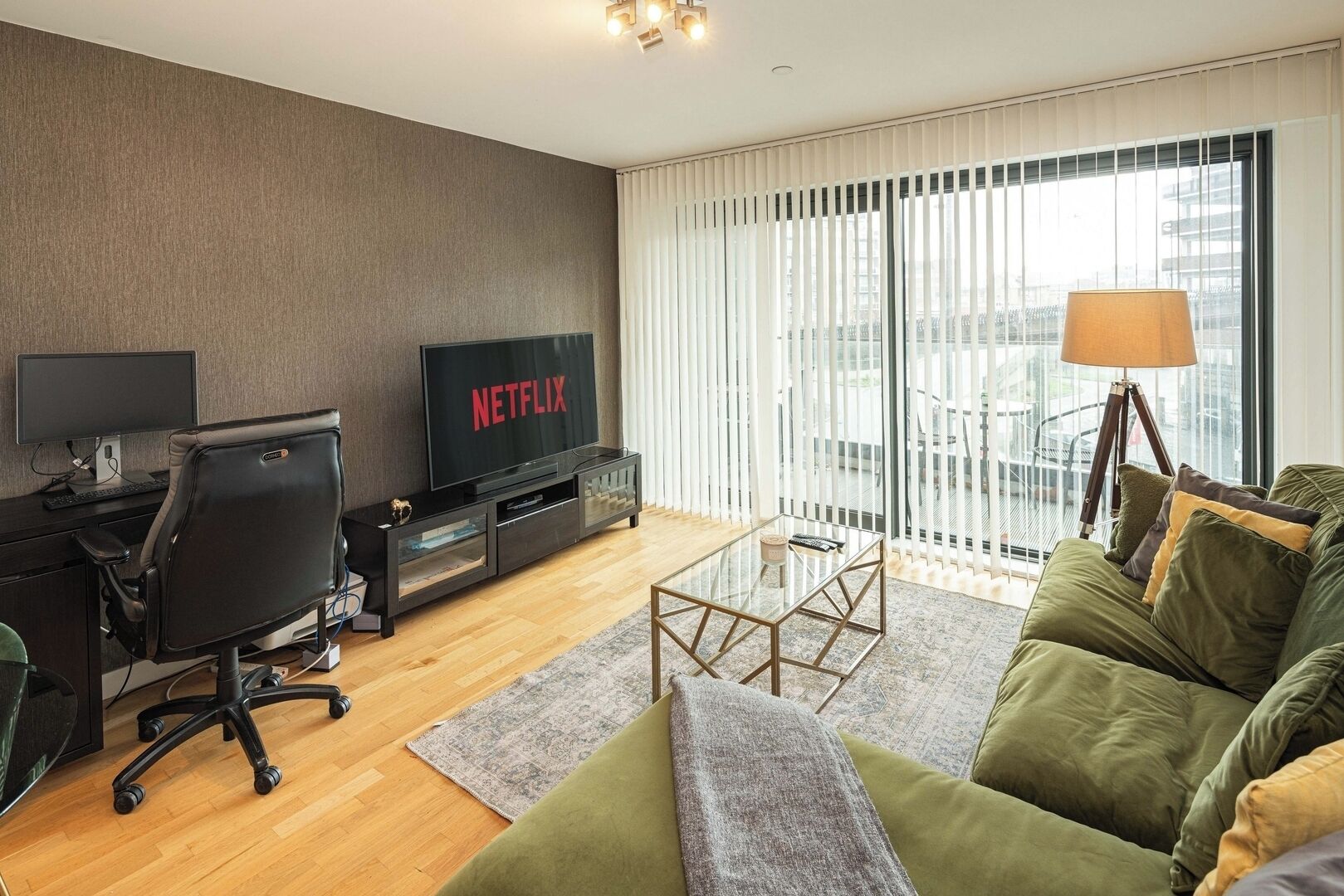 A modern living room with a TV displaying 'NETFLIX', a black office chair, a glass coffee table, and a green sofa with cushions.