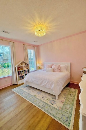 Quarto com cama branca, um candelabro, uma estante de livros e vista para o verde lá fora.