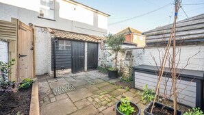 Een achtertuin met een geplaveid terras, een houten poort, een zwarte garage deur en een kleine tuin met planten in potten.