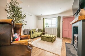 A living room with a leather armchair, a green sofa, a green ottoman, a radiator, a window with curtains, and a pink door. - Pink Cottage, Leiston (Leiston)