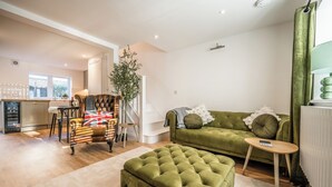 Een zithoek met een groene bank, een gestofte poef, een fauteuil met een patroon en een klein bijzettafeltje. Een plant staat in de hoek, en een raam zorgt voor natuurlijk licht.