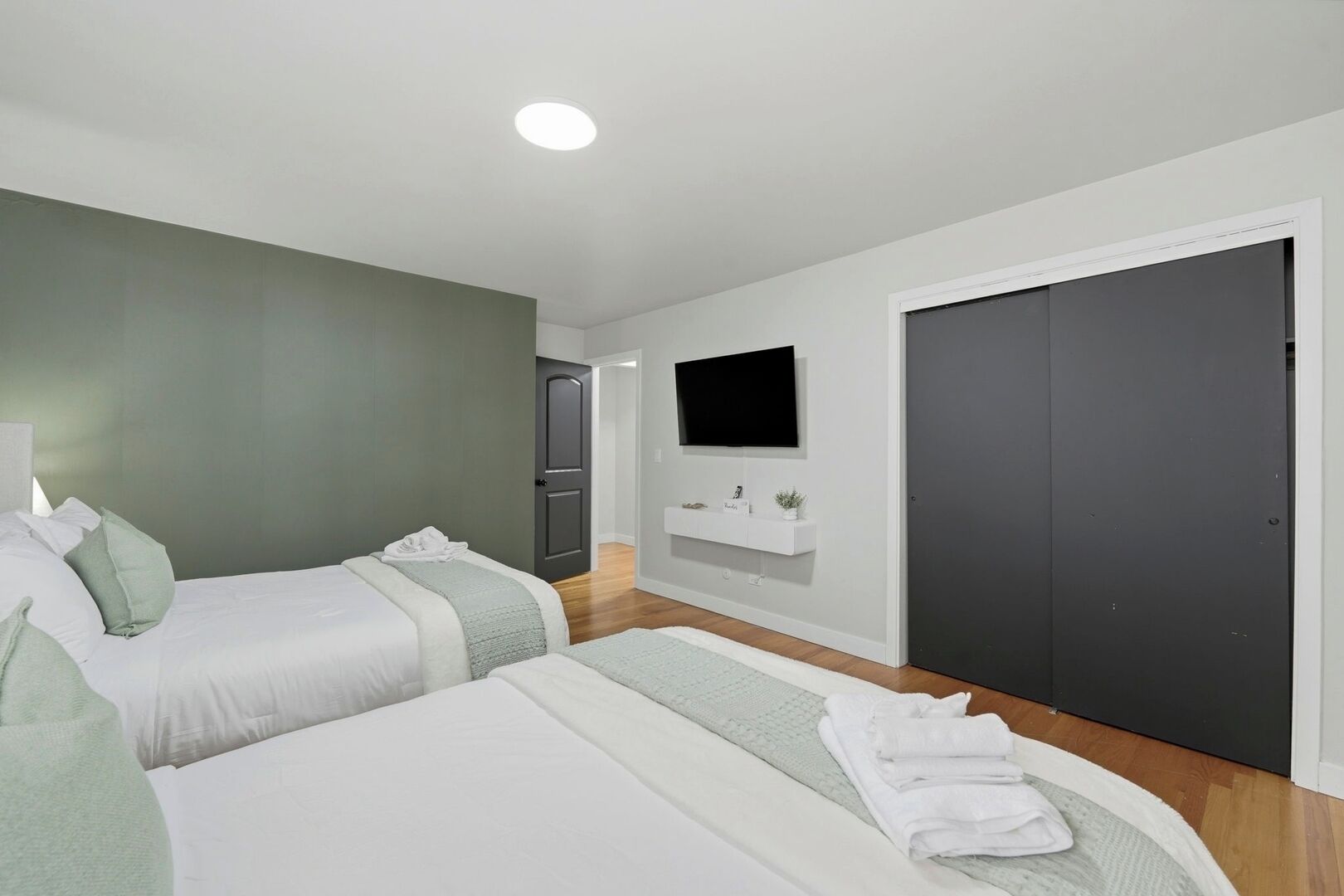 A modern hotel room with two beds, a TV mounted on the wall, a dark door, and a white console table.