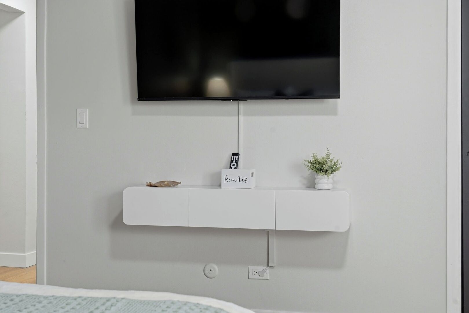 Quarto moderno com televisão na parede, um móvel branco com uma placa, um controle remoto e uma pequena planta.