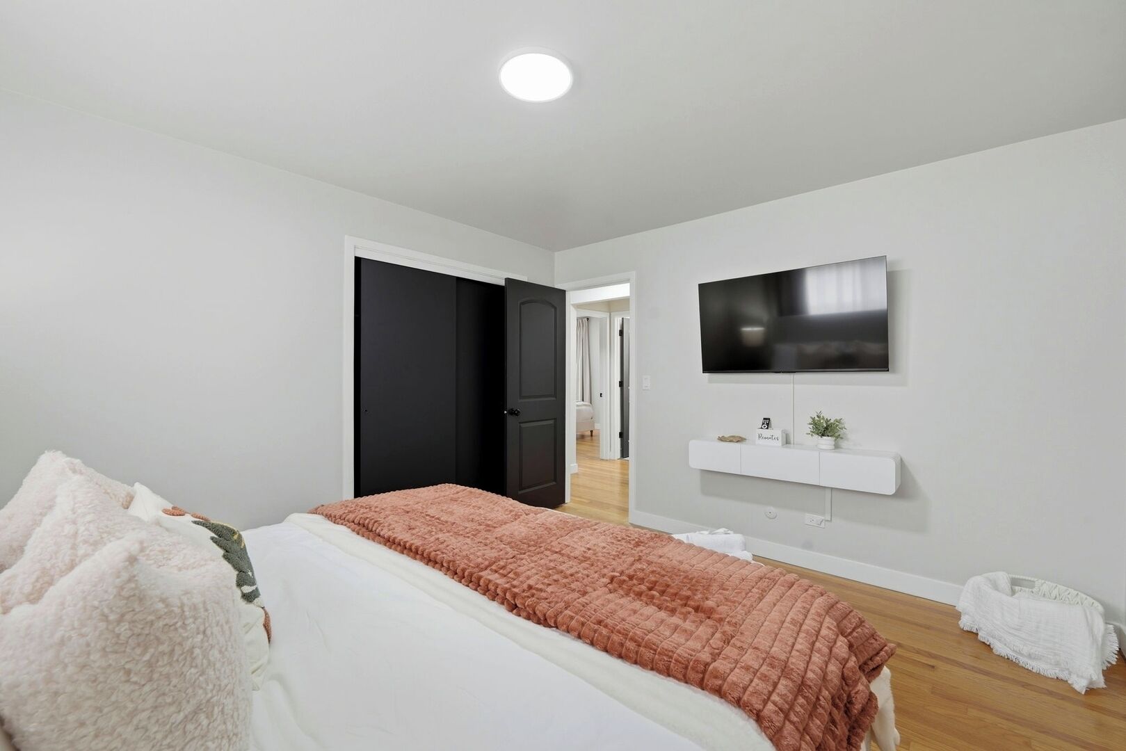 Un dormitorio con cama, televisor, estante blanco con planta y puerta que da a otra habitación.