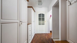 Un couloir aux murs blancs, doté d’un plancher en bois, d’une porte blanche avec un panneau de verre et d’un petit vase contenant des fleurs roses, posé sur une table.