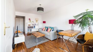 Un salon moderne avec un canapé gris, une table basse en bois et une table à manger avec des chaises. On y trouve également une bibliothèque, une plante en pot et une lampe rouge.