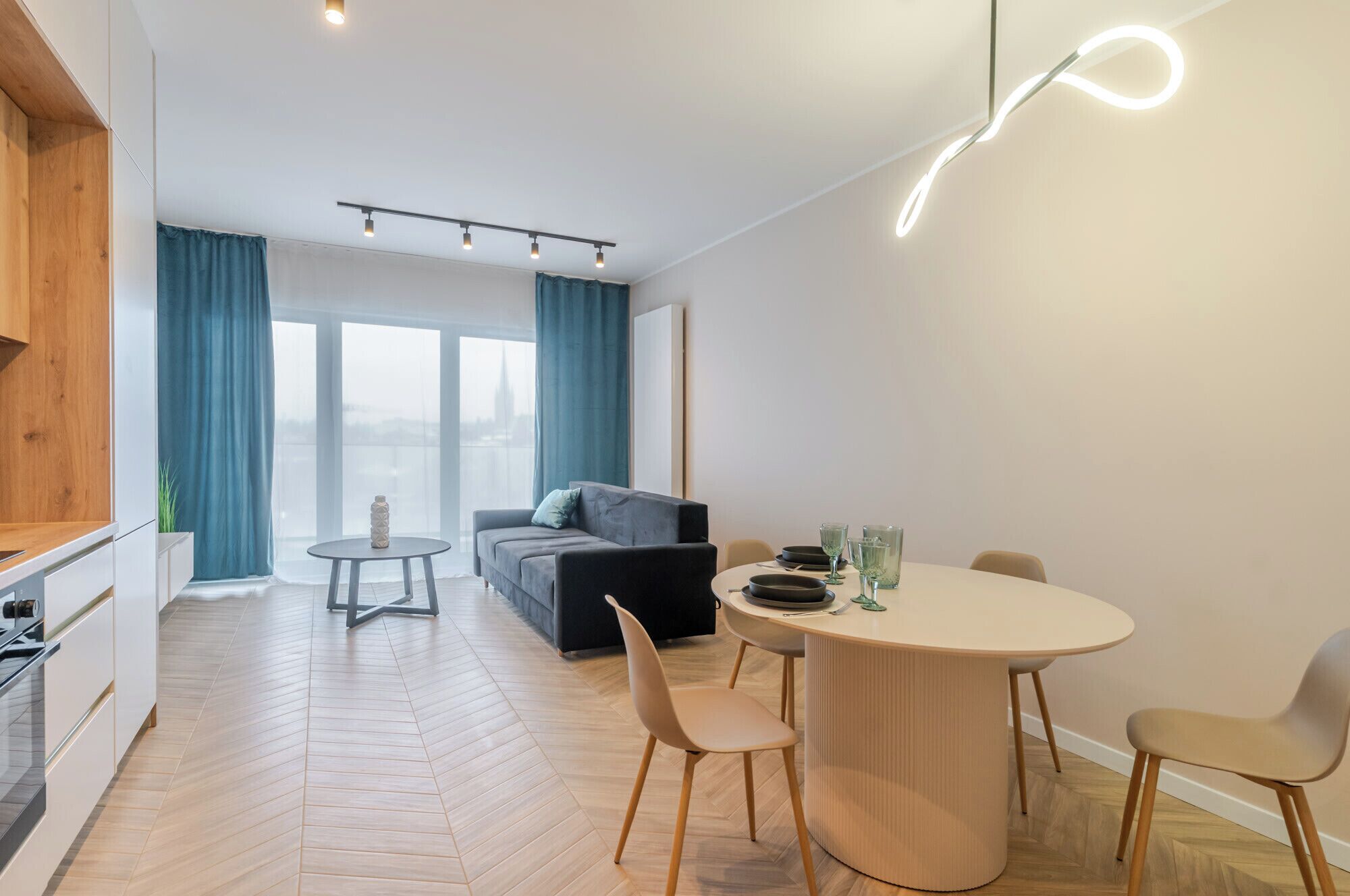 Uma sala de estar moderna com um sofá, uma mesa de jantar com cadeiras, uma mesinha redonda e vista para um horizonte urbano através de grandes janelas.