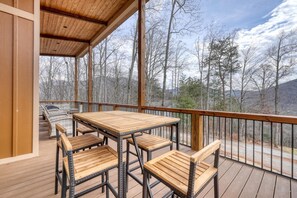 A covered outdoor deck with a table and chairs, overlooking a forested area. - Mossy Oaks Heights - Outdoor Kitchen (Hiawassee)