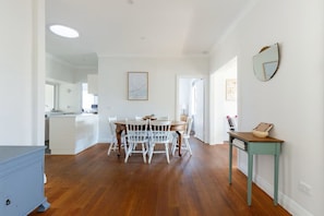 A spacious room with a wooden floor, a dining area with white chairs, a small table with a cushion, and a wall-mounted mirror.